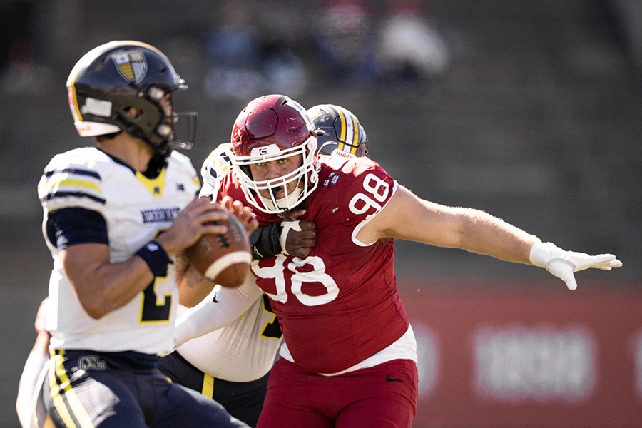 Harvard football player in red jersey 98 rushes Merrimack quarterback in white and blue jersey preparing to throw.
