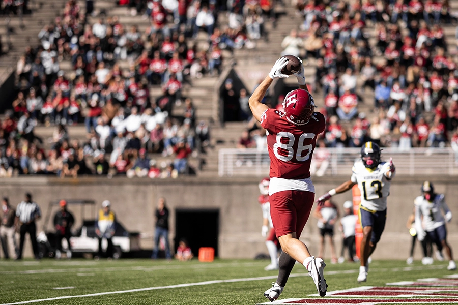 Harvard football player in red jersey 86 catches a football in the end zone with defender trailing behind.