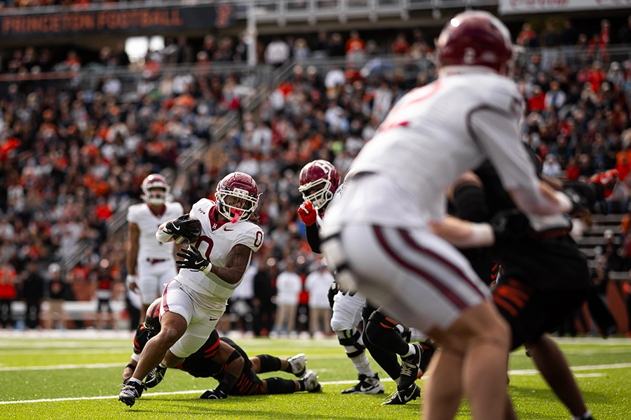 Harvard player runs with football as Princeton defenders attempt a tackle during a game.
