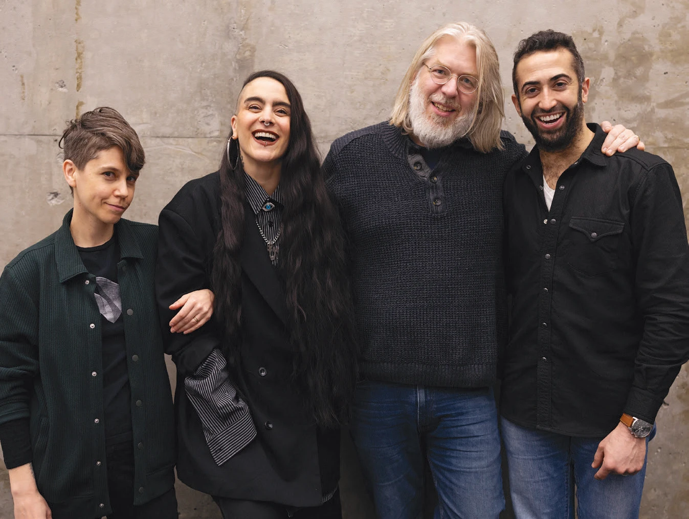 (left to right) Jen Silverman, Sonya Tayeh, Dave Malloy, and Or Matias smiling and standing close together against a concrete wall, arms around each other.