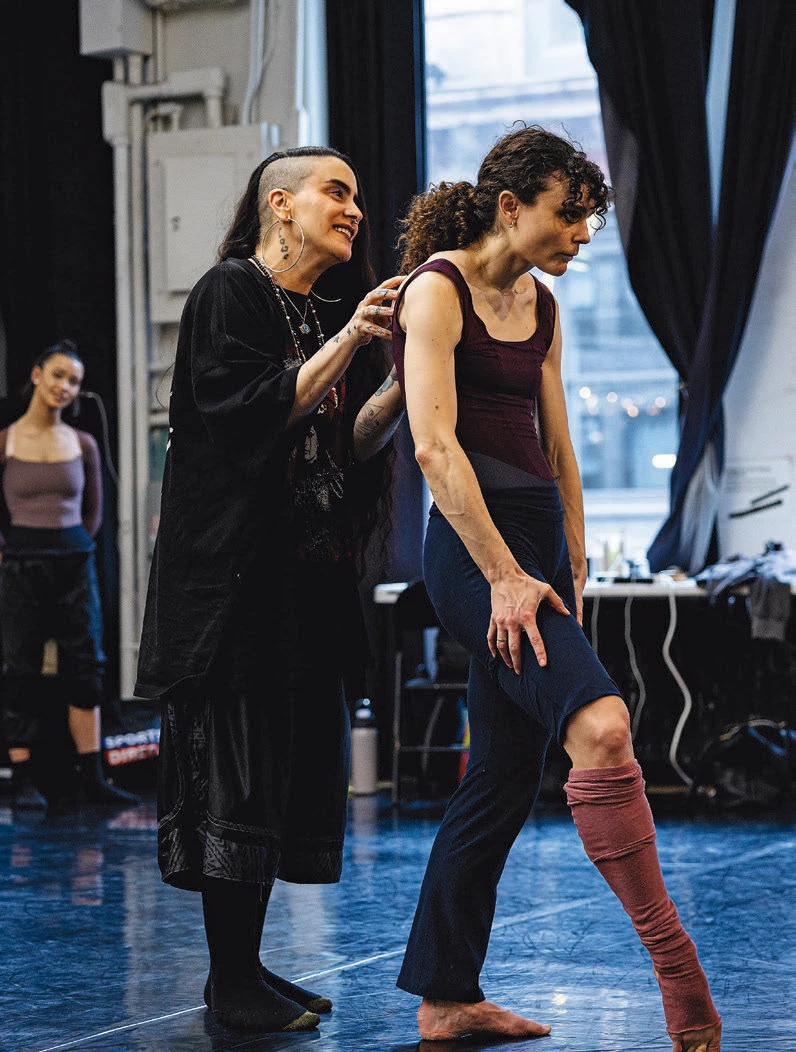 Instructor guiding a dancer’s posture in a studio while another dancer observes in the background.