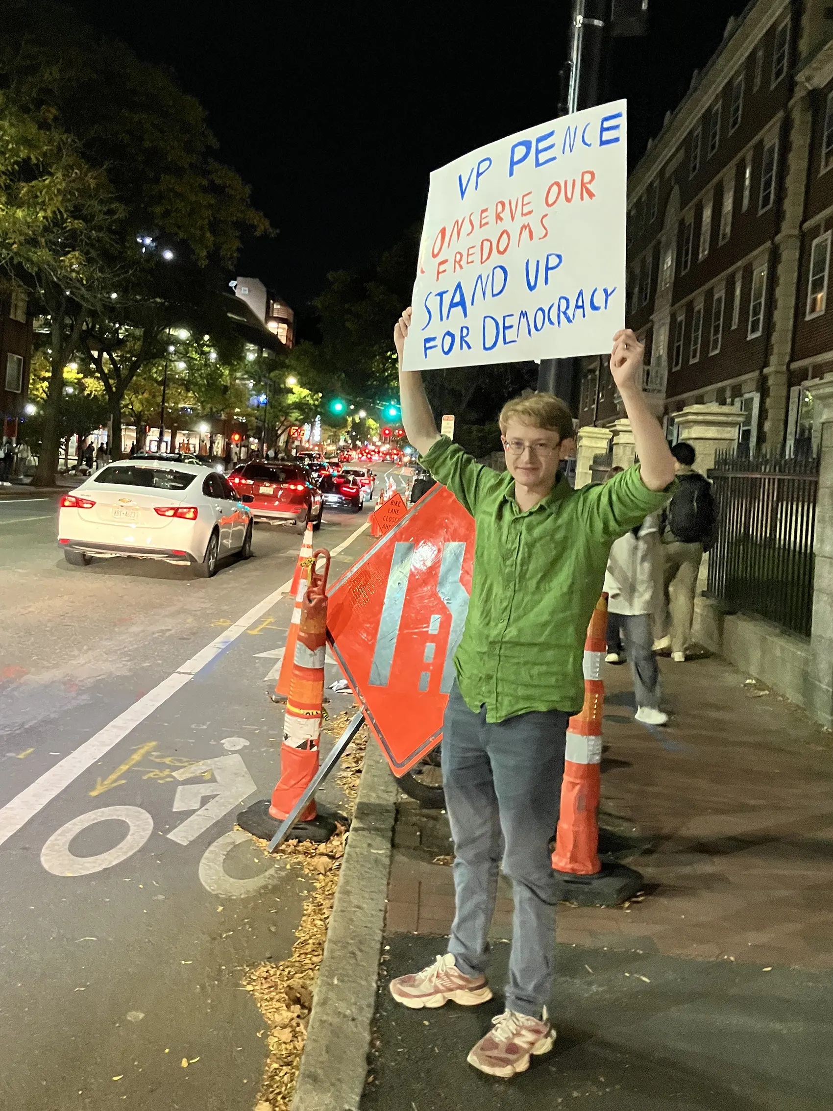 Harvard student protests outside of Kennedy School, holding sign reading "VP Pence Conserve our Freedoms Stand Up for Democracy"