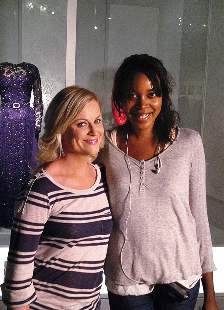 Aisha Muharrar with Amy Poehler smiling on the Parks and Rec set with a sequined dress in the background.