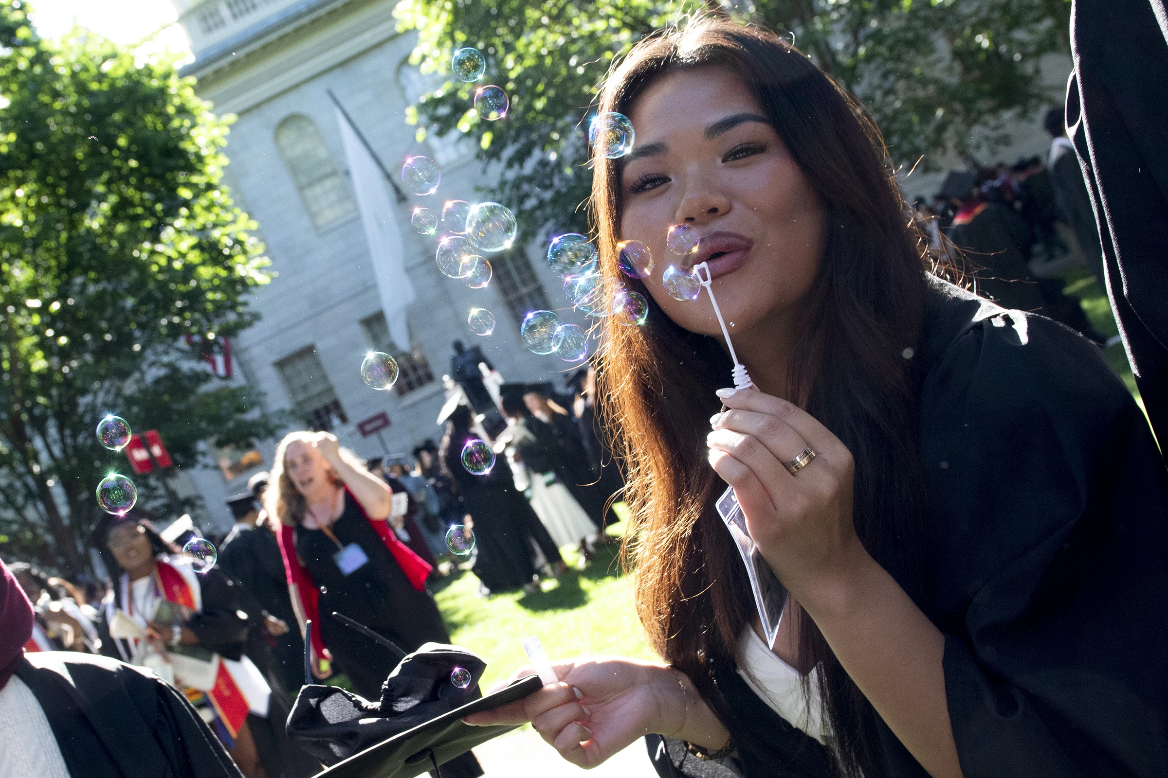 Woman wearing black robe and holding bubble blower, blowing bubbles in front of white brick building