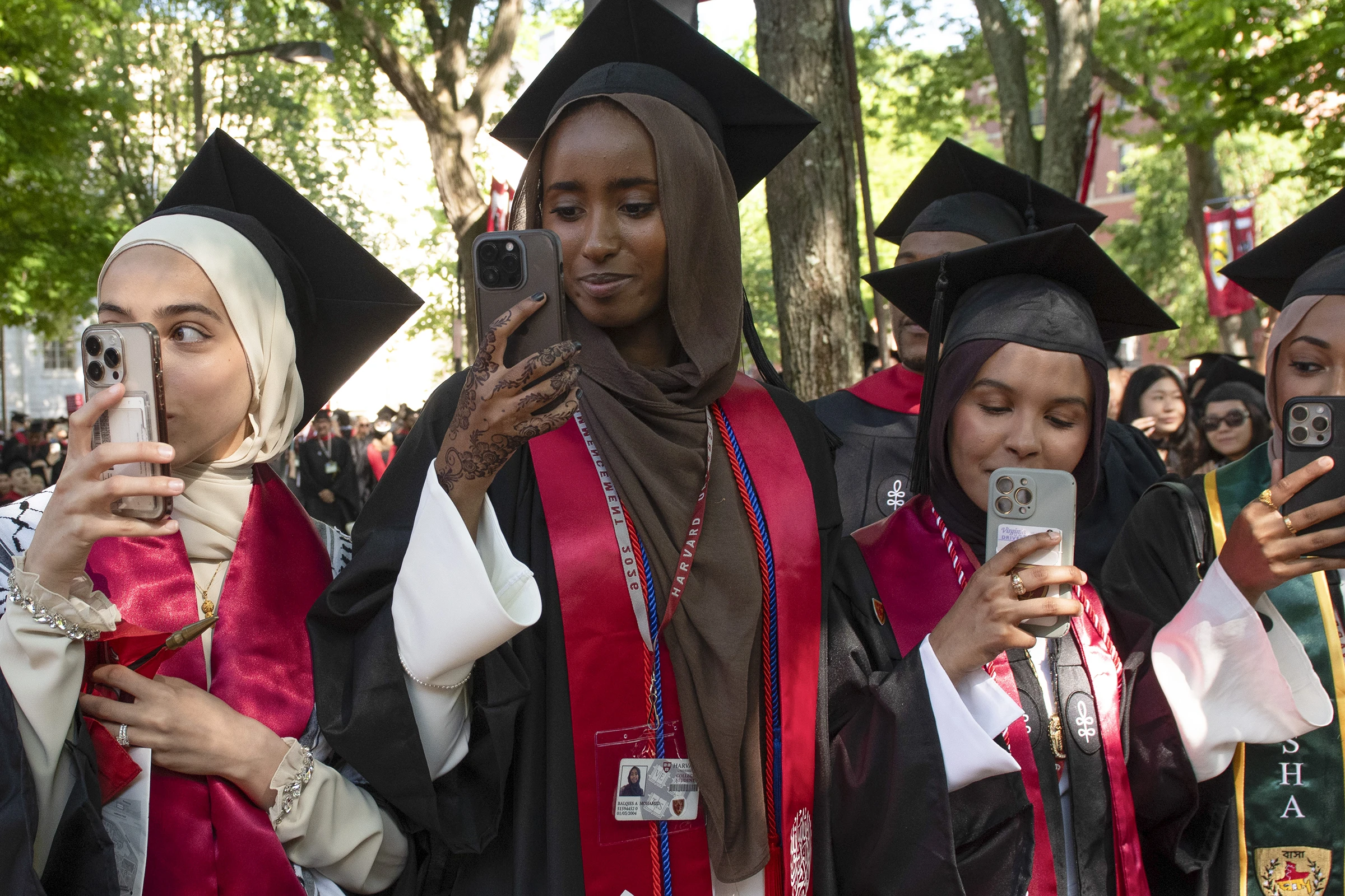 Three women wearing hijab and black mortarboard hats with red stoles holding up their phones to take a photo with trees in the background