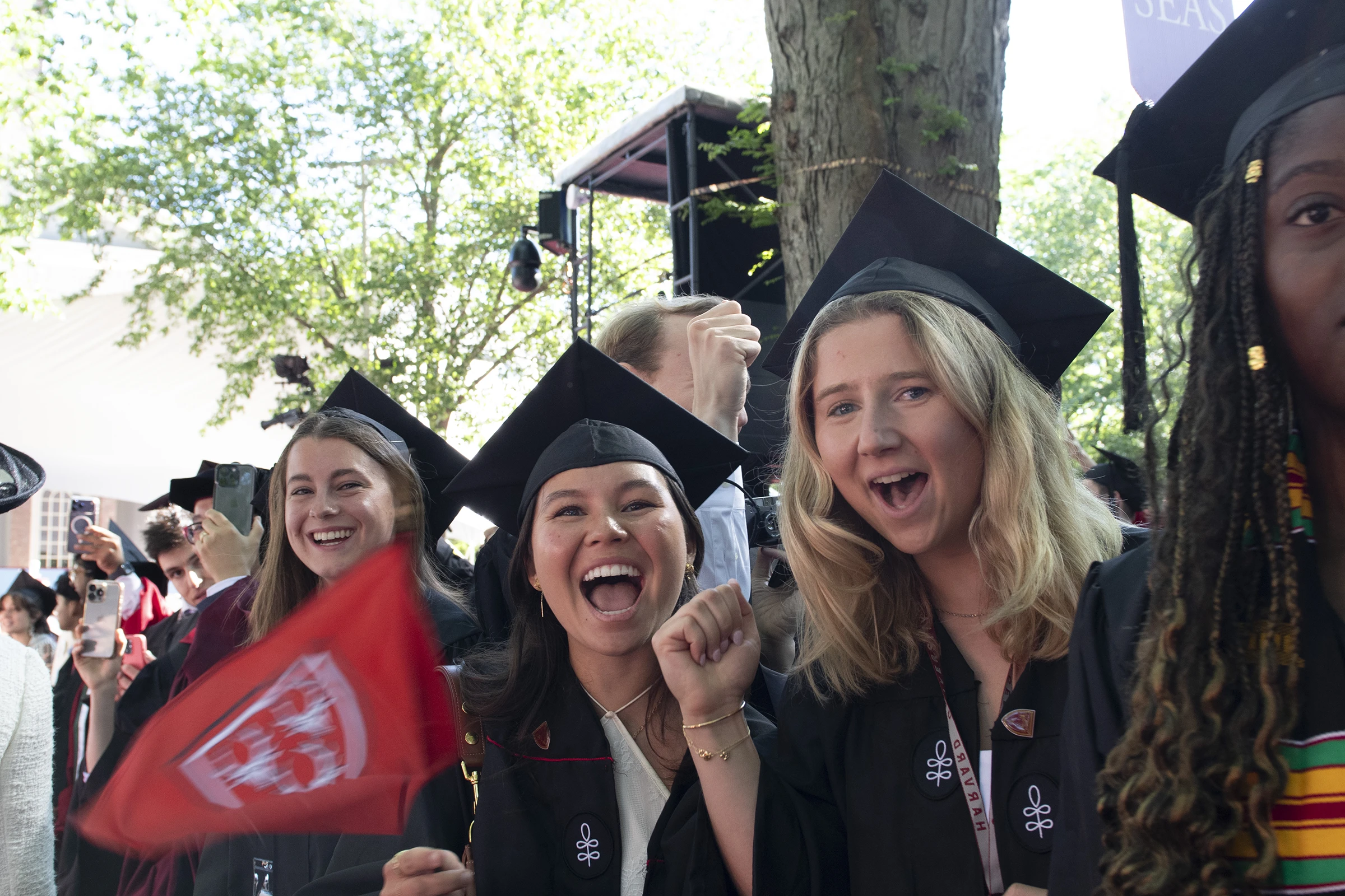 Three women wearing black graduation mortarboard caps cheer and wave a red flag 