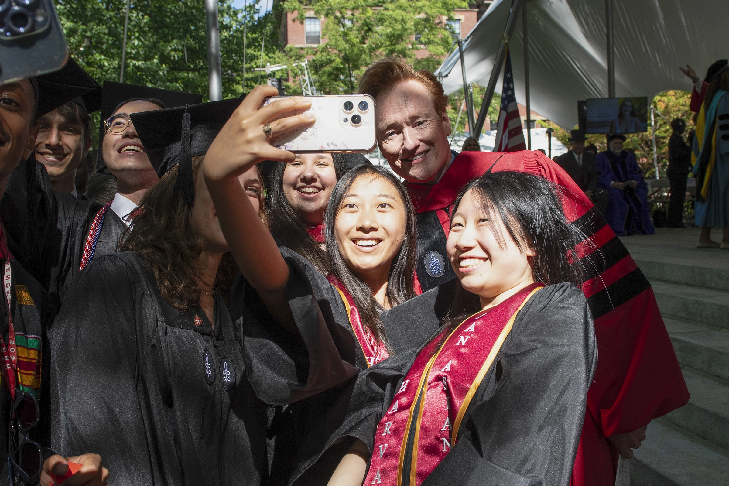 Students standing in a crowd wearing black graduation caps and gowns holding up a phone, taking a photo with a man with red hair and red gown