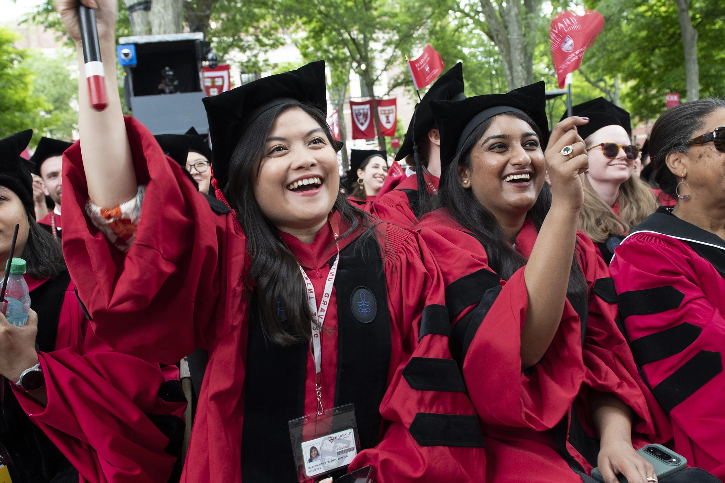 Students wearing red PhD graduation robes with black stripes and black stoles smile and wave red flags in the air with trees and red banners in the background