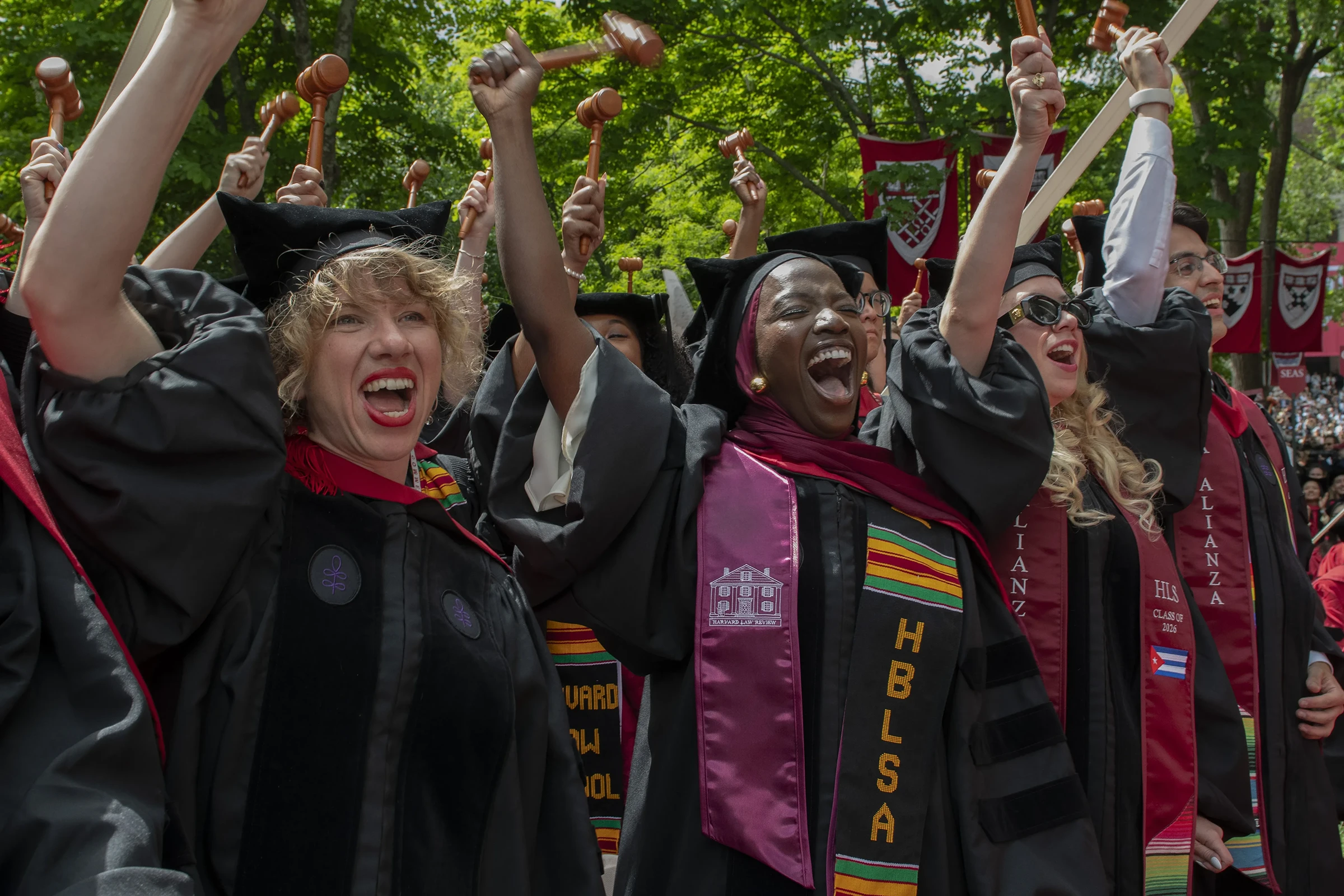 Students wearing black graduation caps and gowns cheer with mouths open and holding gavels