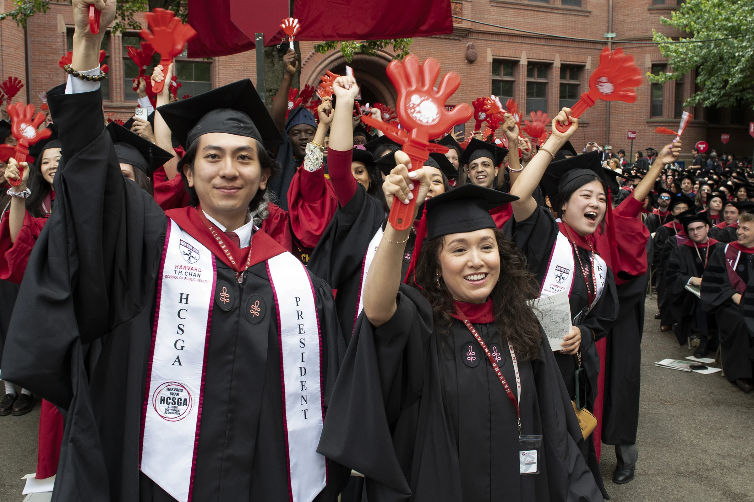 Students wearing black graduation gowns and caps hold up red plastic clapper hands