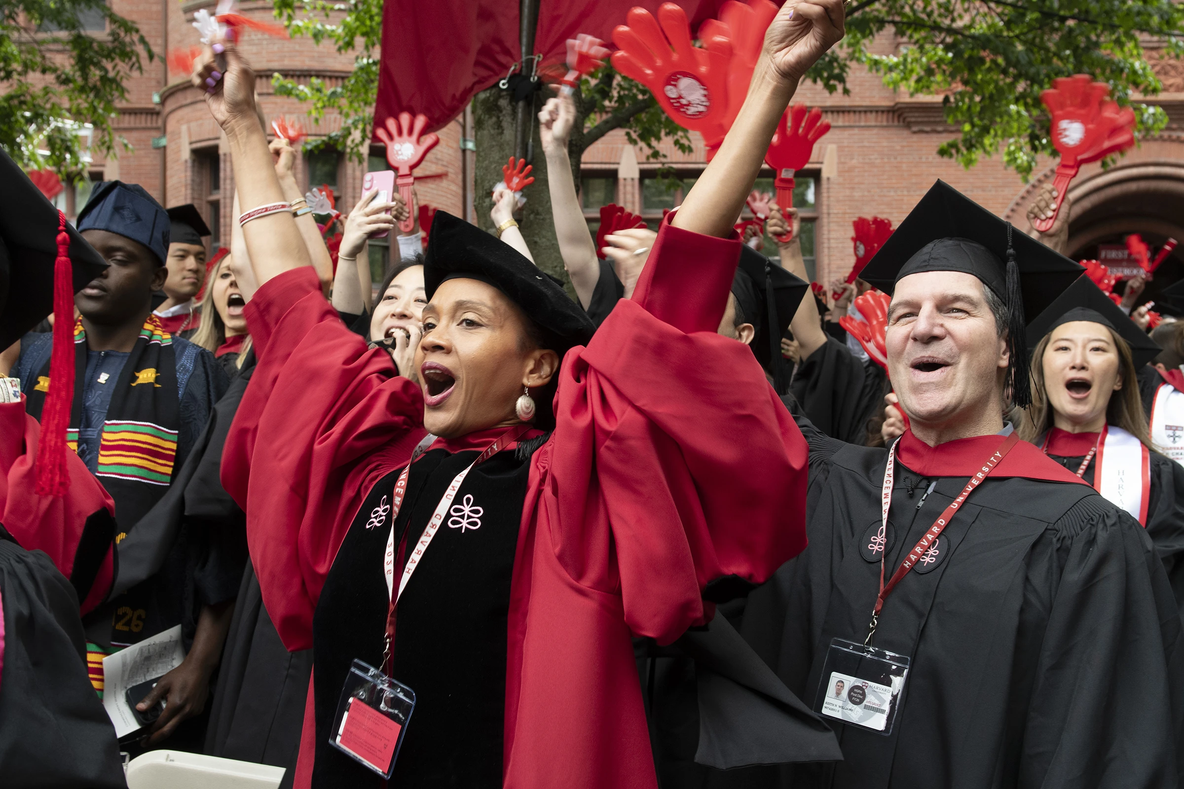 Students wearing red and black robes and black graduation caps cheer with arms up, holding red plastic clapper hand toys