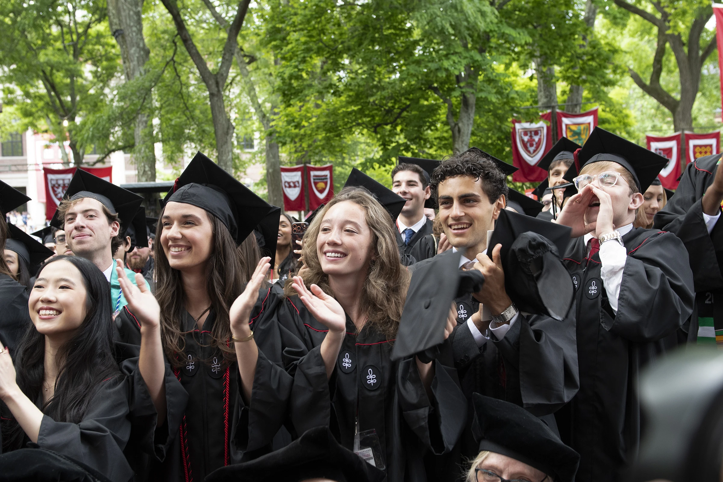 Students in black graduation cap and gowns clap with red banners and trees in the background
