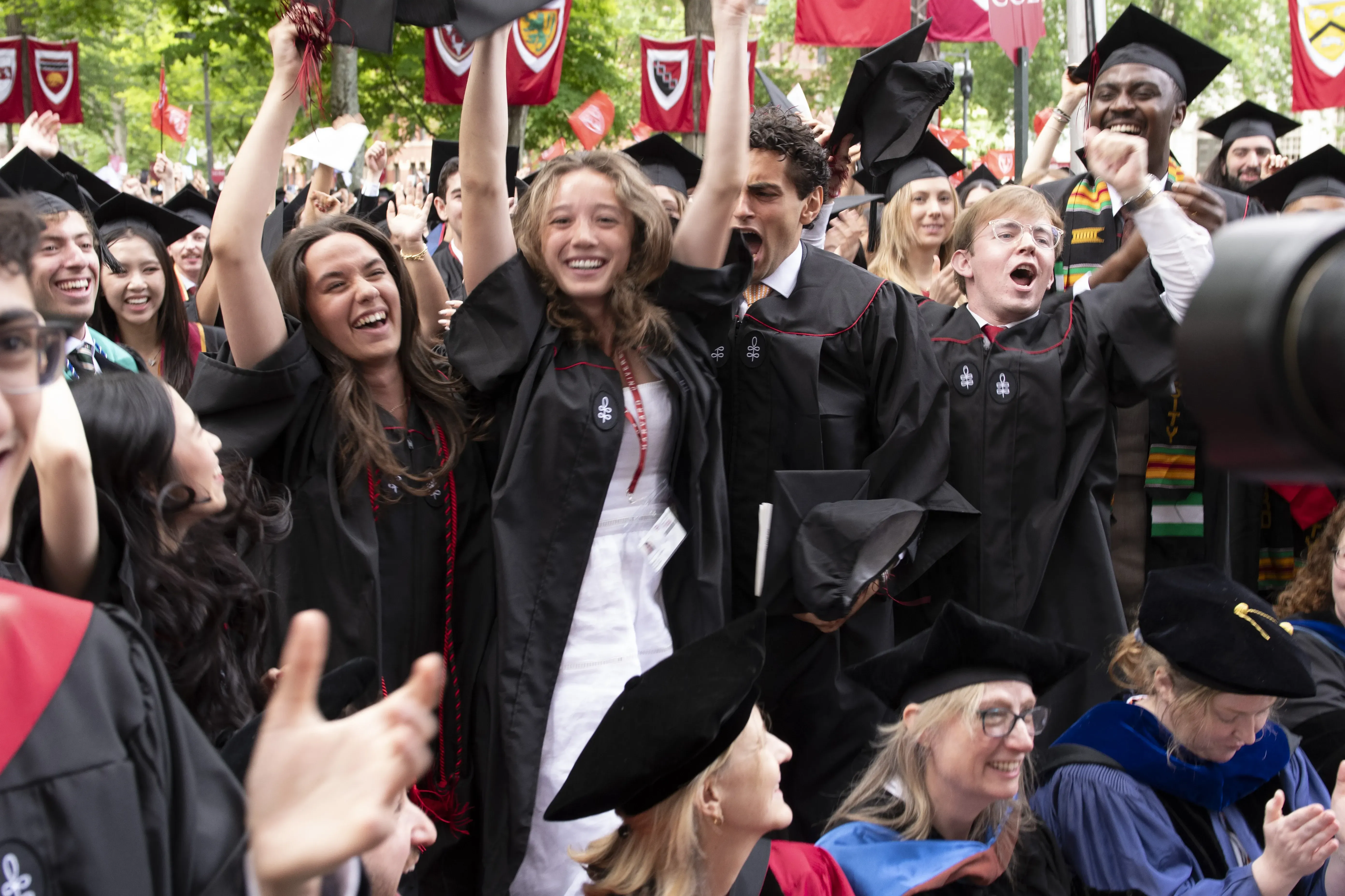 Students in black robes and black graduation caps cheering and jumping in the air with red banners in the background