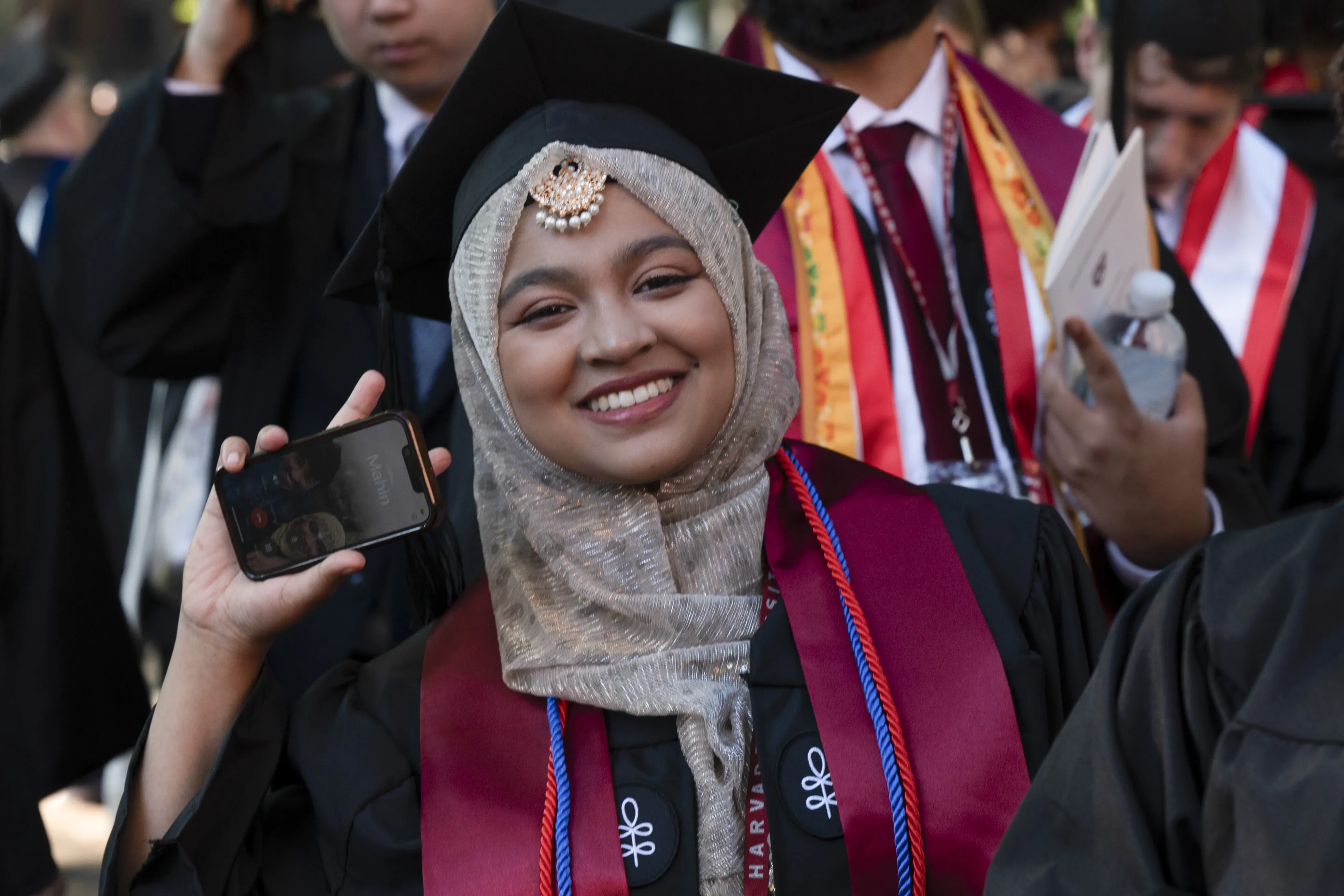 Woman wearing beige hijab with pears at the top and a black mortarboard cap smiling and holding a phone