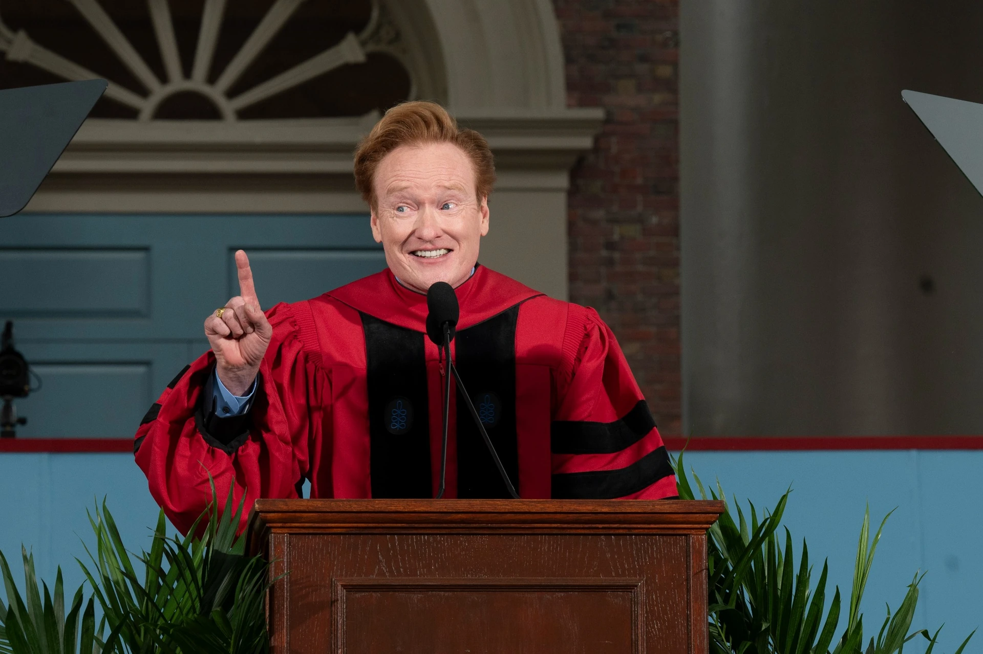 Conan O'Brien smiles and points his finger up while wearing a red doctoral robe and speaking from a podium
