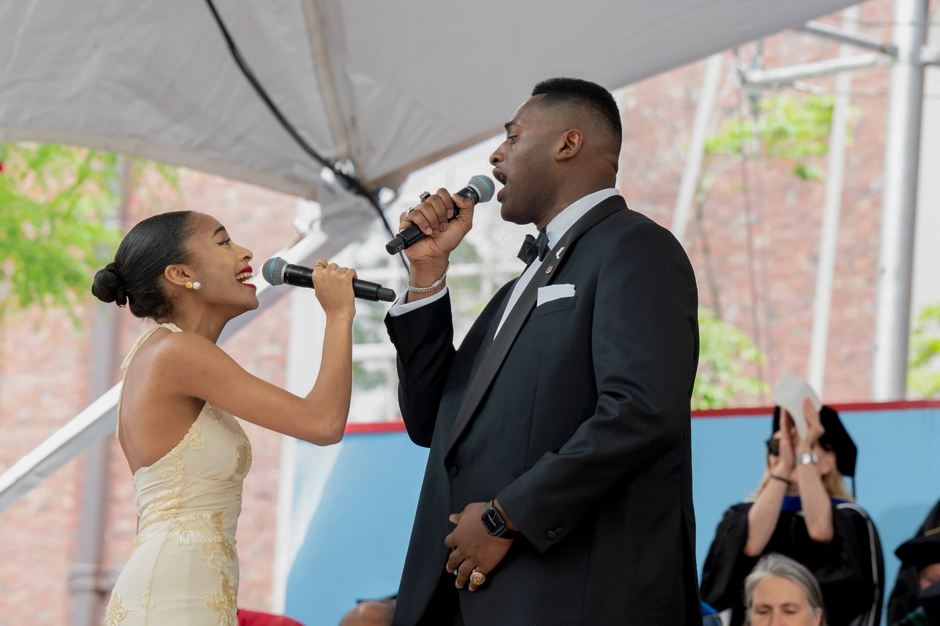 Female singer wearing gold gown and male singer wearing black tuxedo sing to each other holding microphones 
