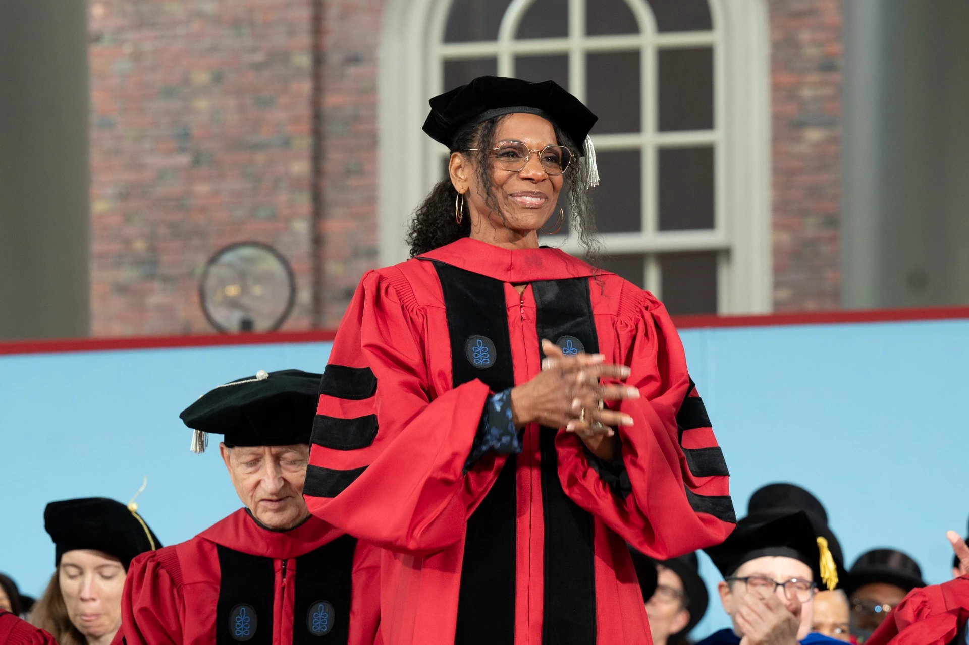 Audra McDonald smiling and wearing a red doctoral robe stands on a stage with hands clasped 