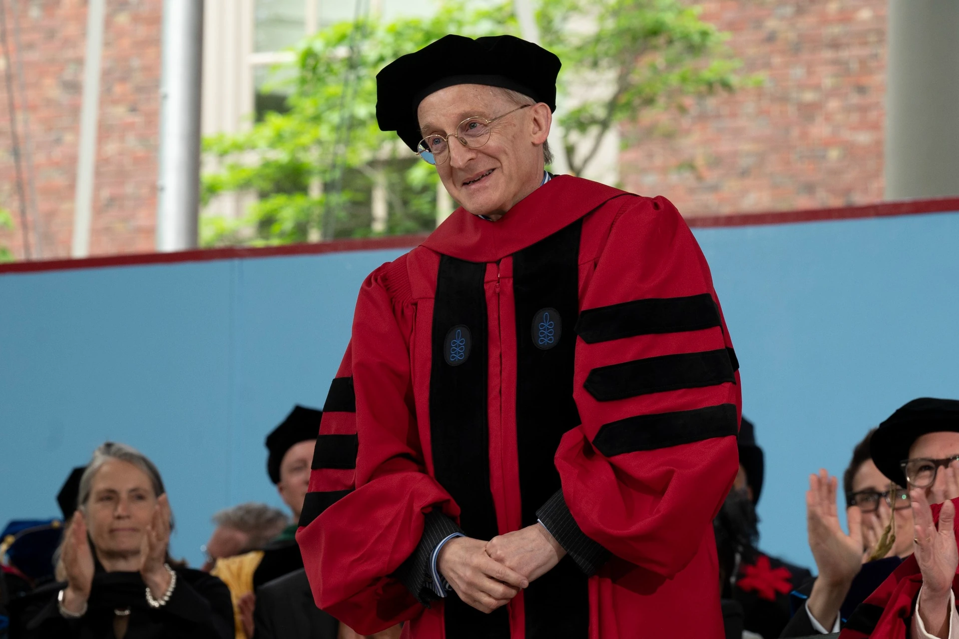 Sir Noel Malcolm smiles while wearing a red doctoral robe on a stage