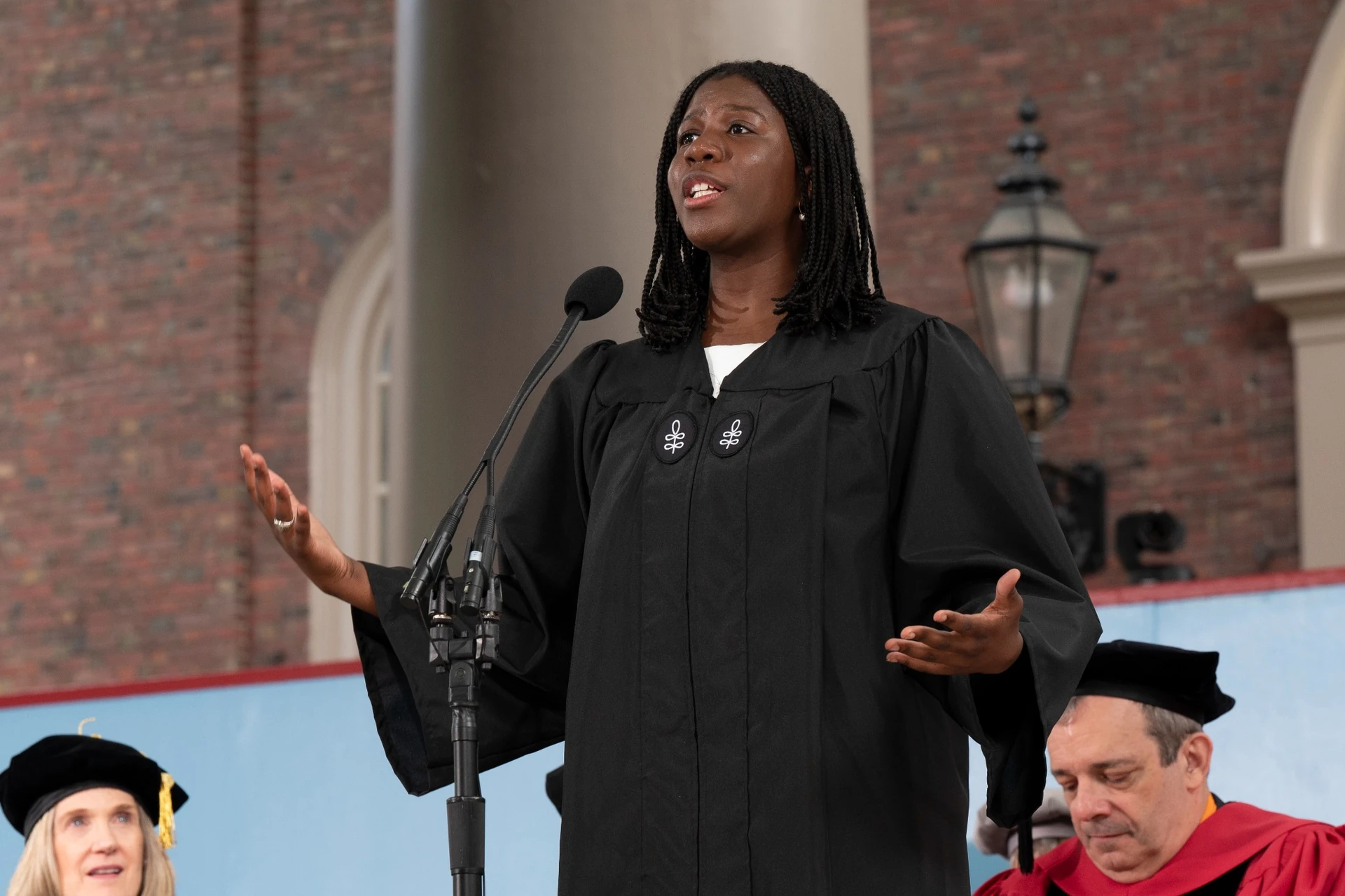young woman in black robe with arms raised delivers a speech on a stage