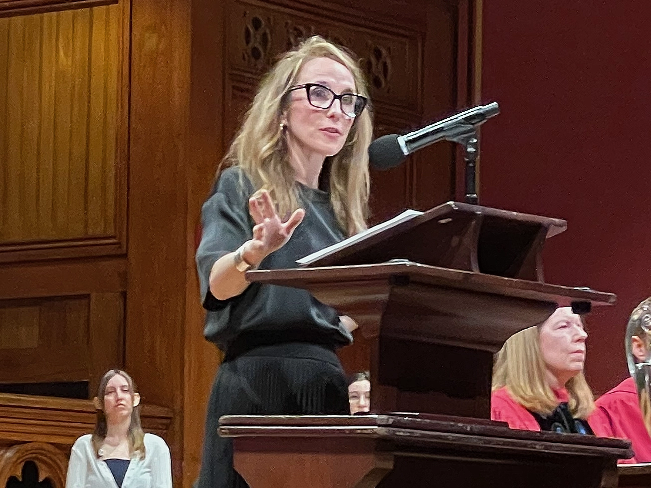 Meghan O’Rourke wearing a black dress and black eyeglasses with long blond hair stands at the podium speaking