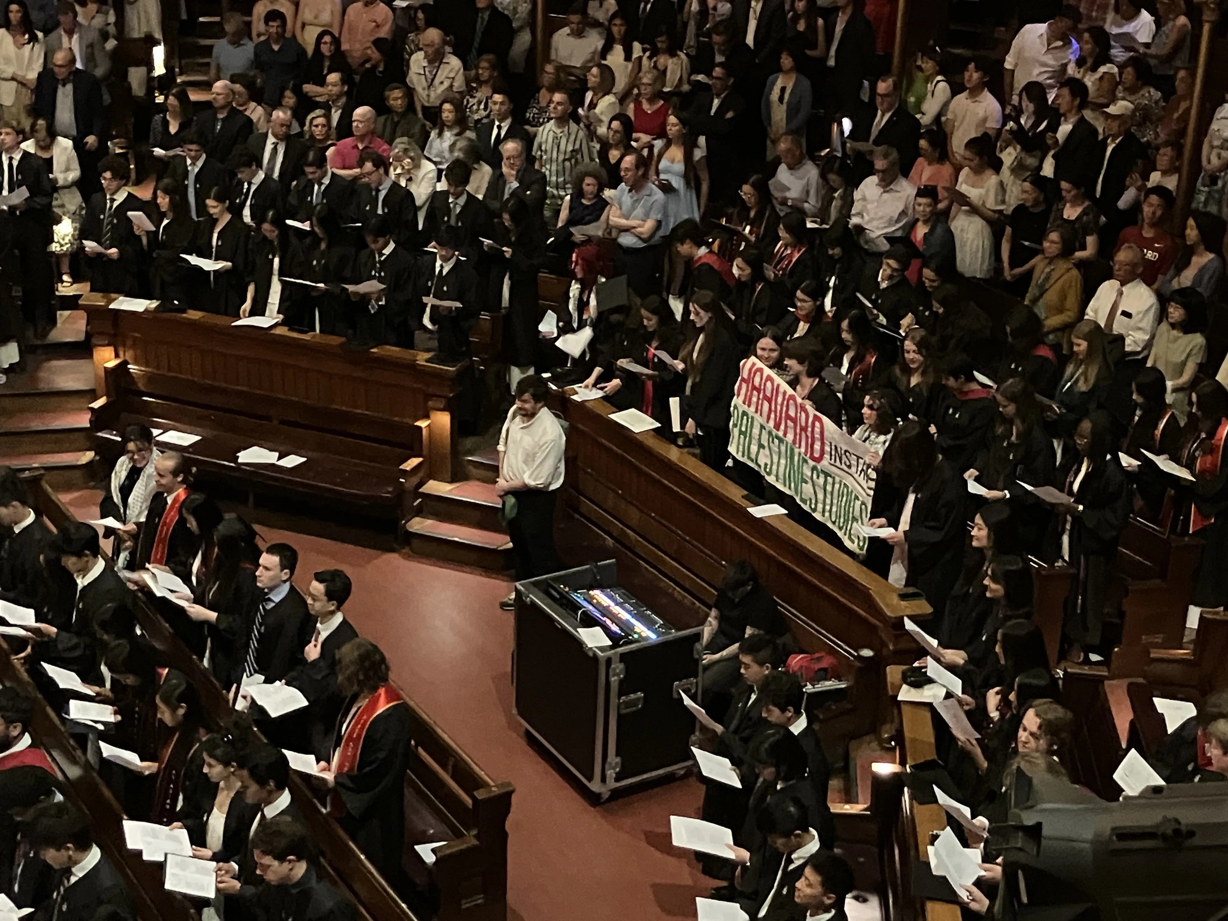 Crowd of students standing holding papers in an auditorium with three students holding up banner that reads "Harvard Instate Palestine Studies"