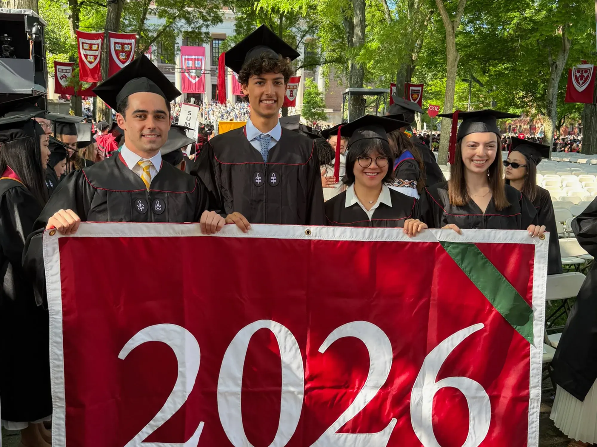 Four graduates in black cap and gown hold a red banner with "2026" on it