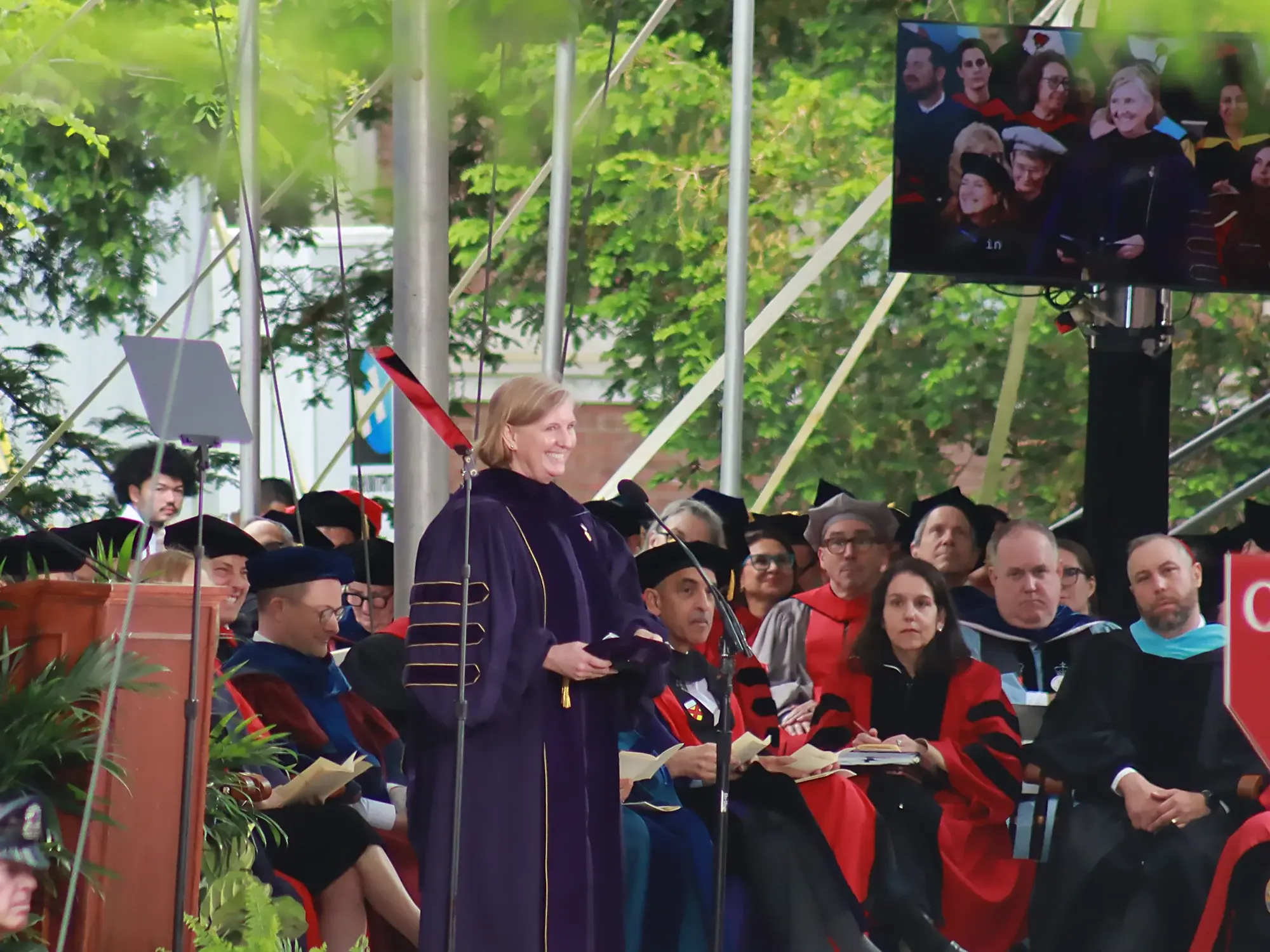 Woman in purple and black graduation gown standing at microphone speaking to a crowd with people in red gowns and black caps sitting behind her on stage