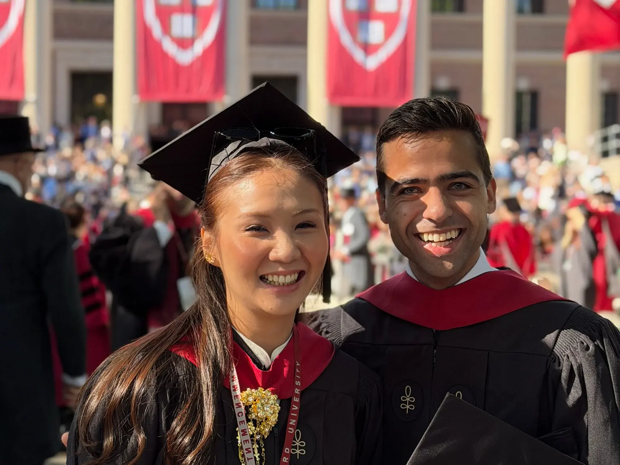 A woman in a graduation mortarboard cap smiling with a man wearing a black graduation robe with pillars and red banners in the background