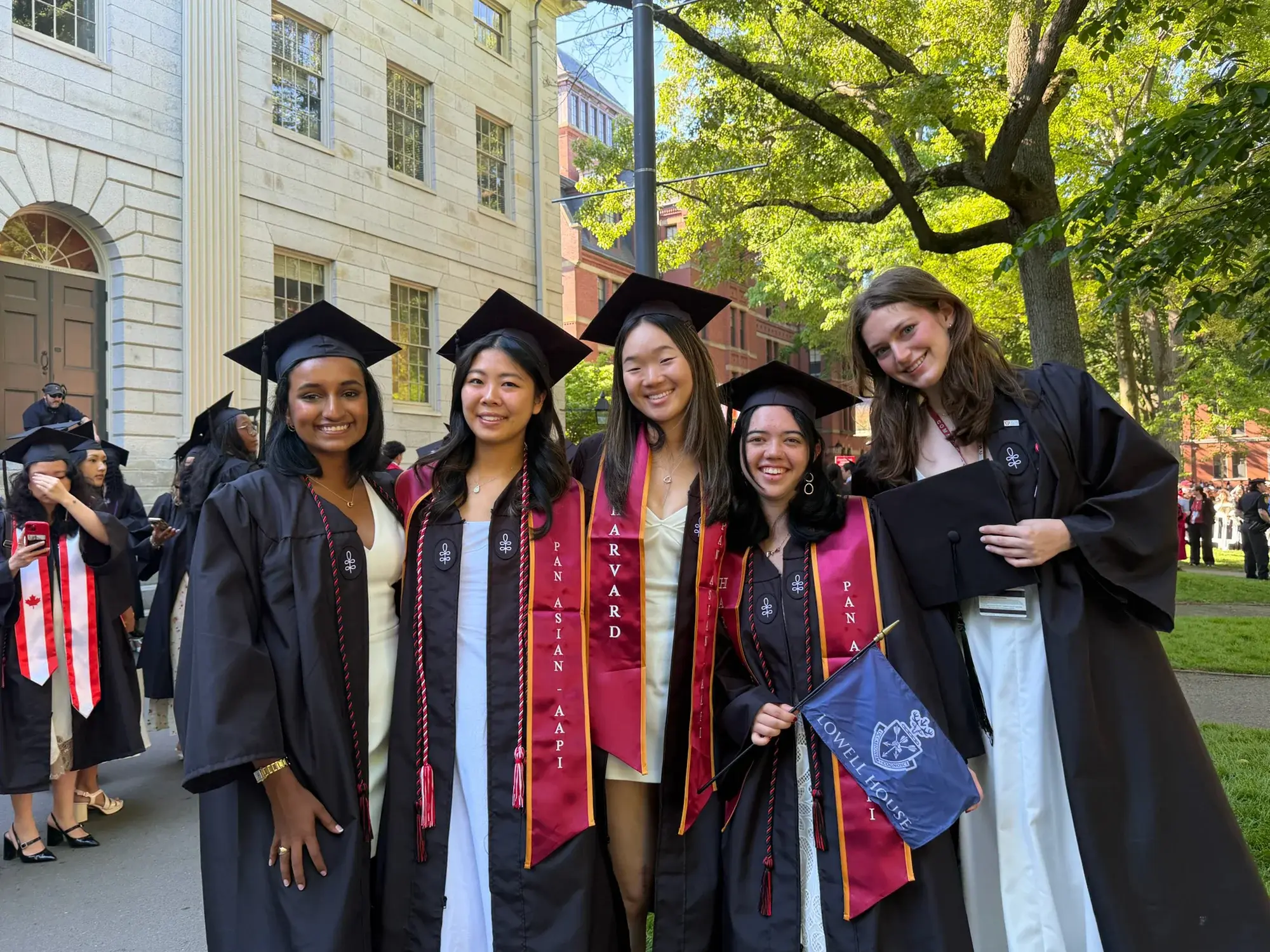 Four women wearing black mortarboard caps and gowns with red stoles, holding a blue flag with a crest and "Lowell House"