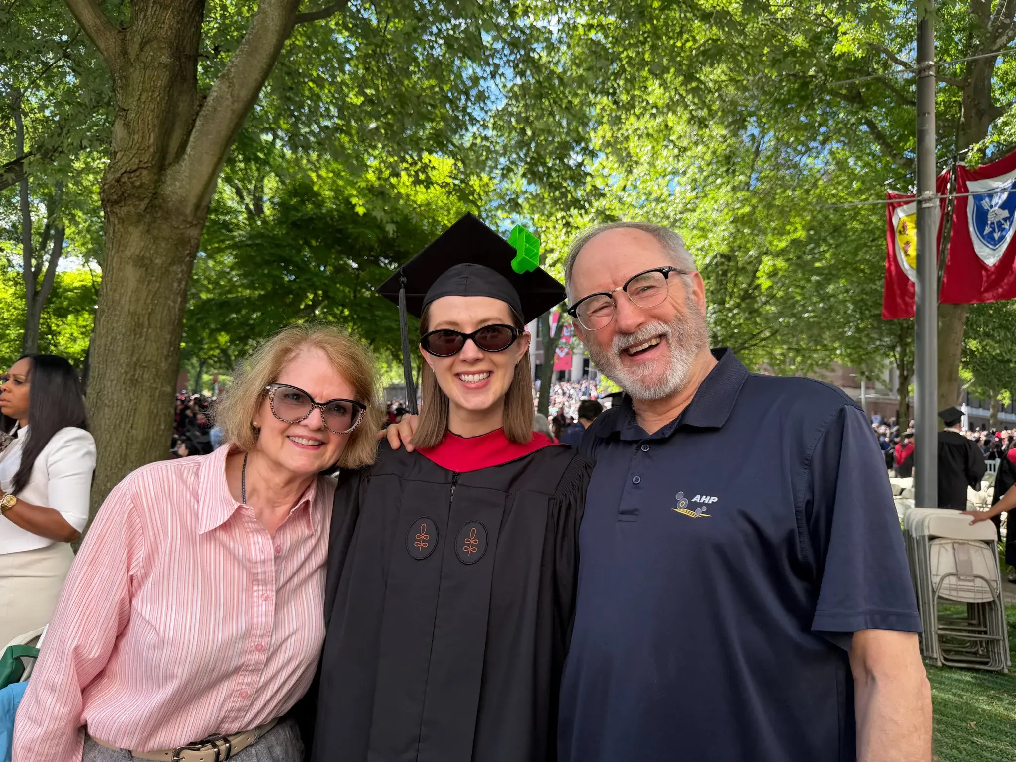 Woman with sunglasses and pink collared shirt, woman wearing sunglasses and black graduation gown and mortarboard cap, man wearing glasses and navy polo shirt, with trees in the background