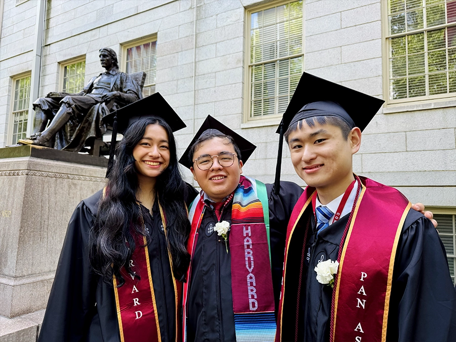 Harvard graduates wearing regalia gather near the John Harvard sign