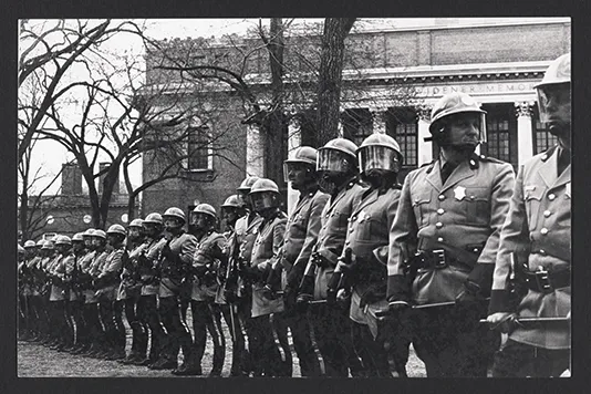Black-and-white photo of a line of uniformed police in riot gear standing outside Harvard’s Widener Library during a protest.
