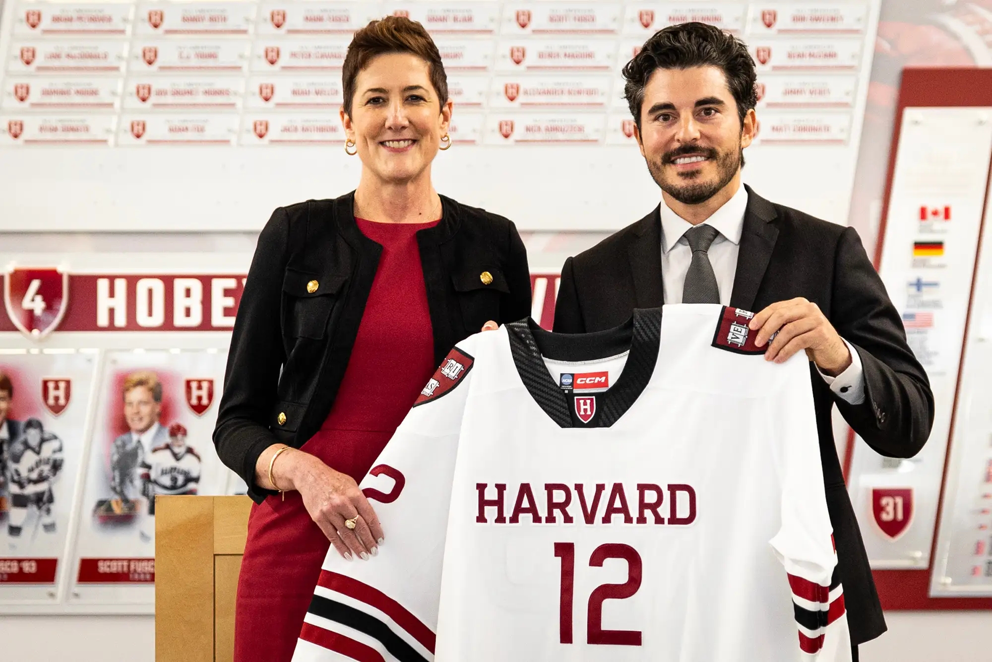 Two people in business attire hold a white Harvard hockey jersey with number 12 during a presentation in front of team displays.