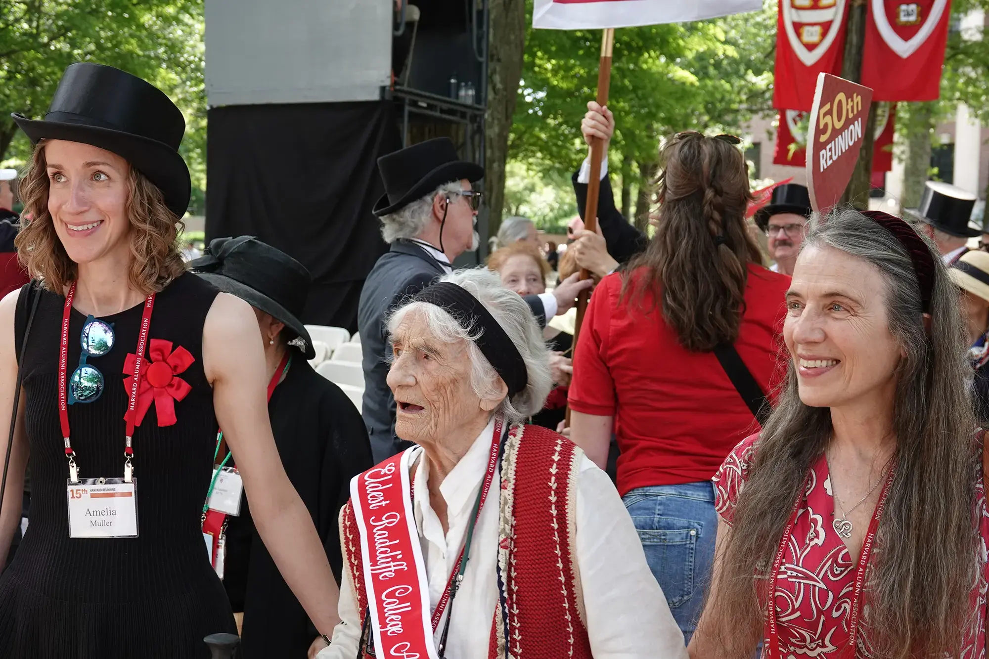 a woman wtih a black top hat and red ribbon, an elderly woman with a red "Eldest Radcliffe College" sash, a woman with a black headband and red dress