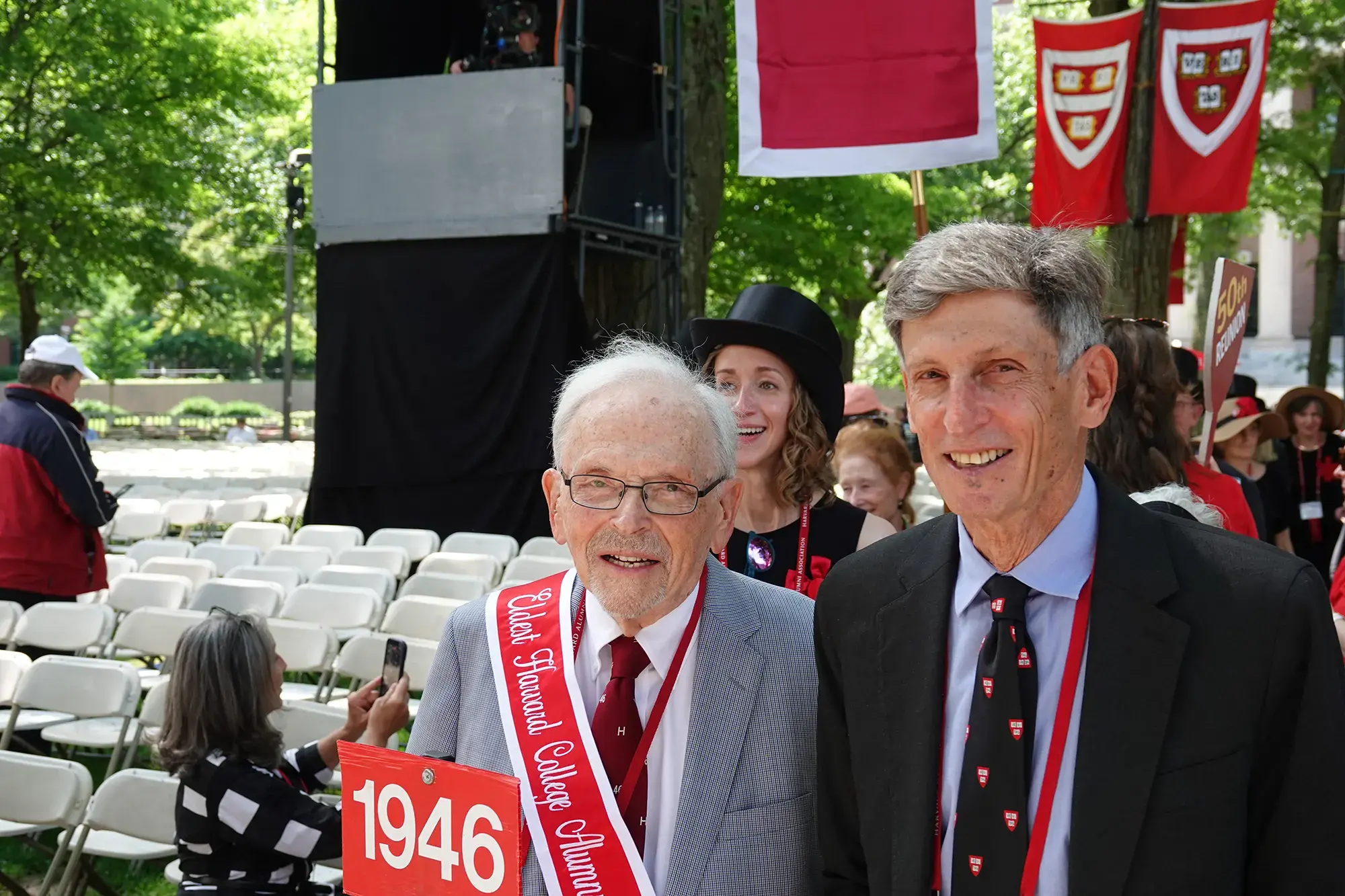An elderly man holding a red "1946" sign and a red "Eldest Harvard College Alumni" sash, standing next to a man with a black tie with Harvard shields on it