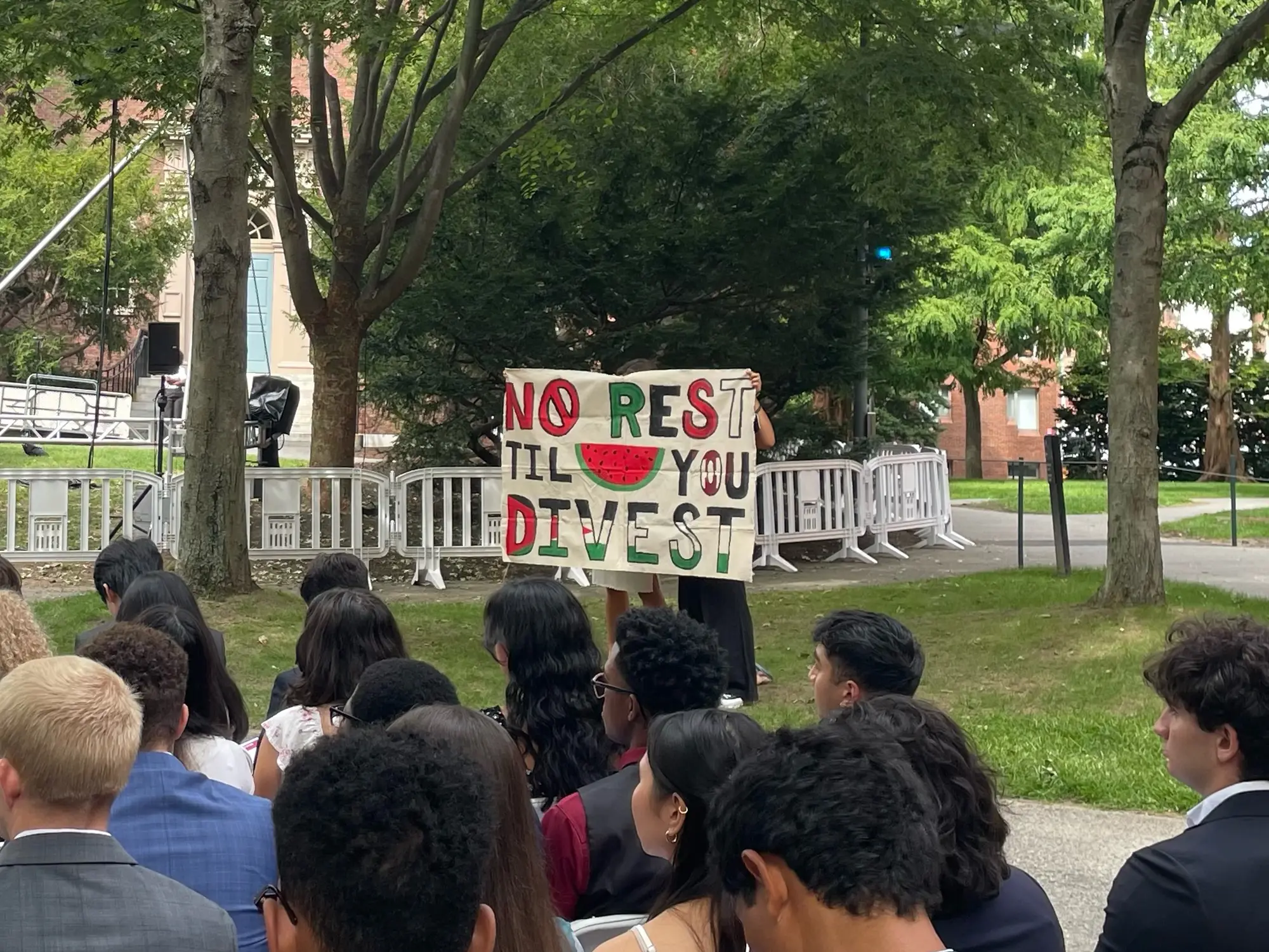 Hand painted protest sign in Tercentenary Theatre that reads "No Rest Til You Divest" with a watermelon graphic in the middle held up by two people. Students sit in the foreground