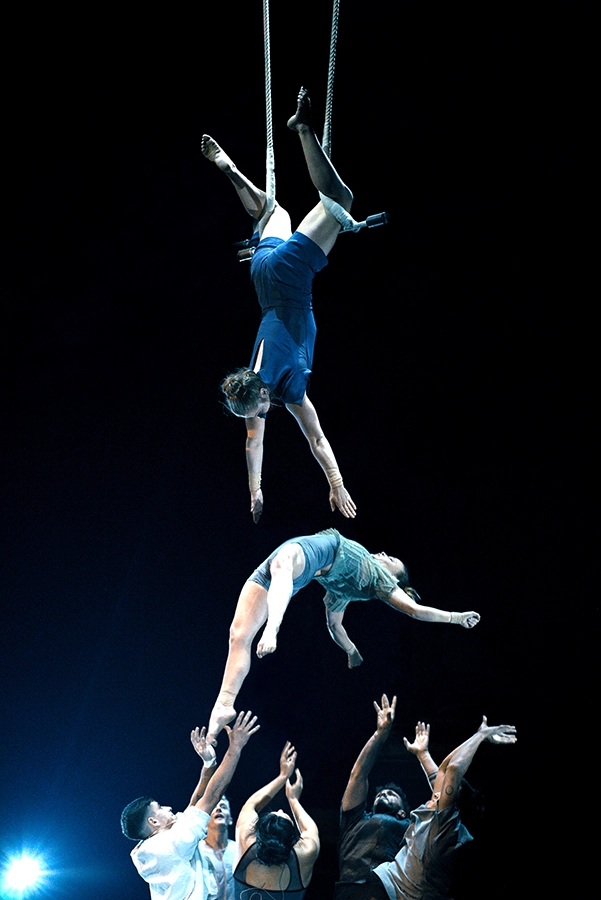 aA performer hangs upside down on straps while another acrobat is midair above outstretched arms.