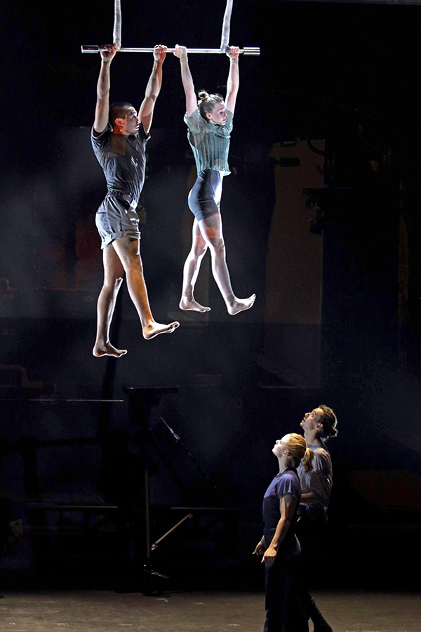 Two aerial performers hang from a trapeze as two others watch from below.