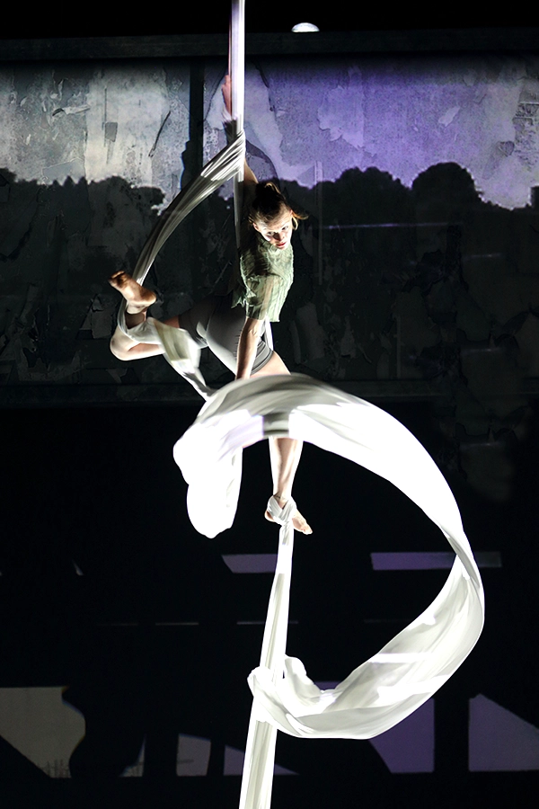 Aerialist poses midair wrapped in flowing white silks against a dark backdrop.