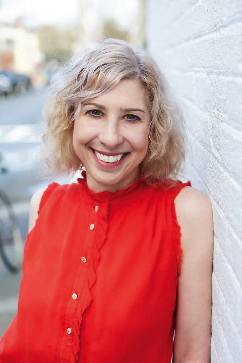 Joanna Weiss, wearing a red shirt, standing against a white brick wall