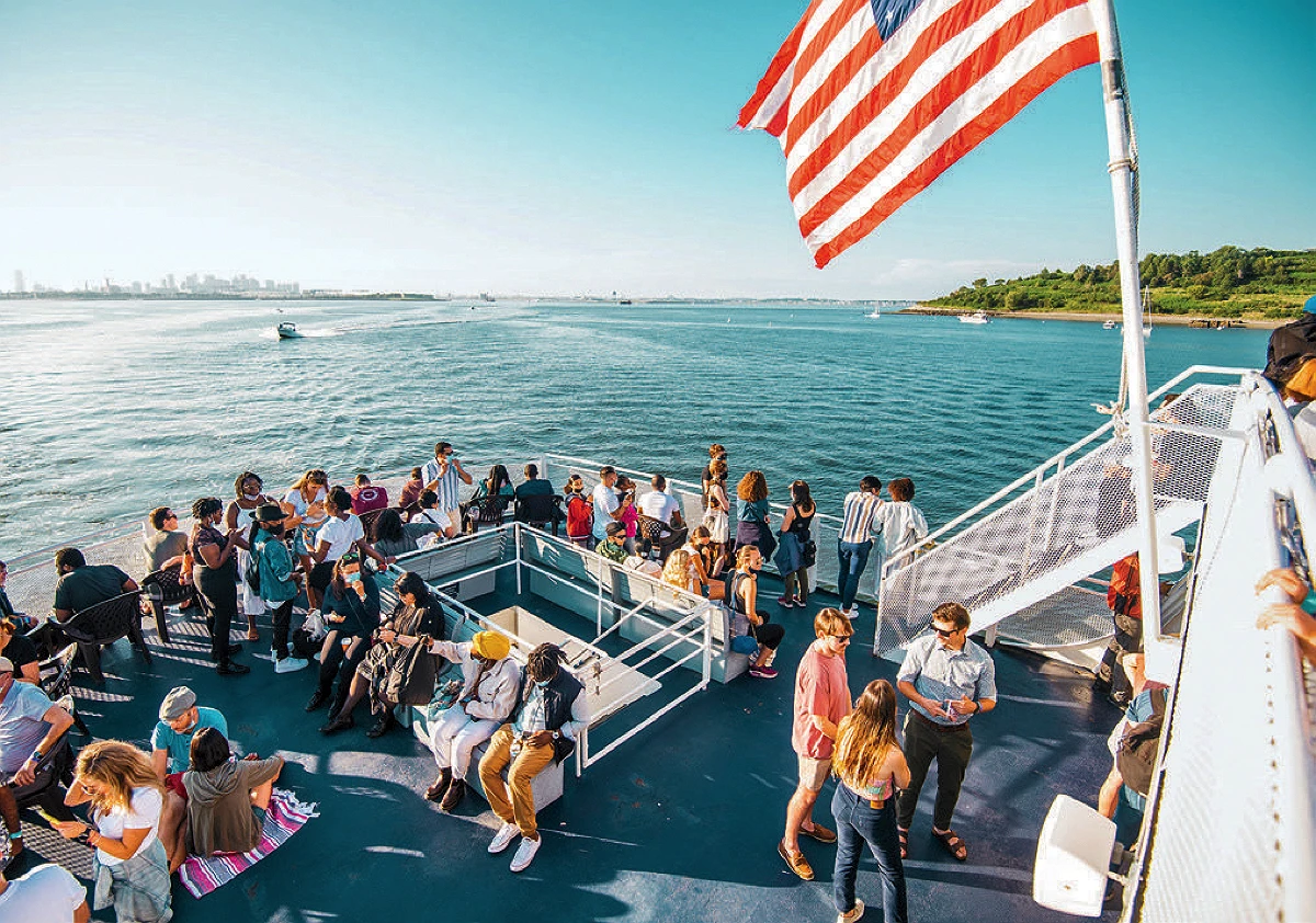 A diverse group of people socializing on a ferry deck under a U.S. flag, with ocean and coastline in the background.