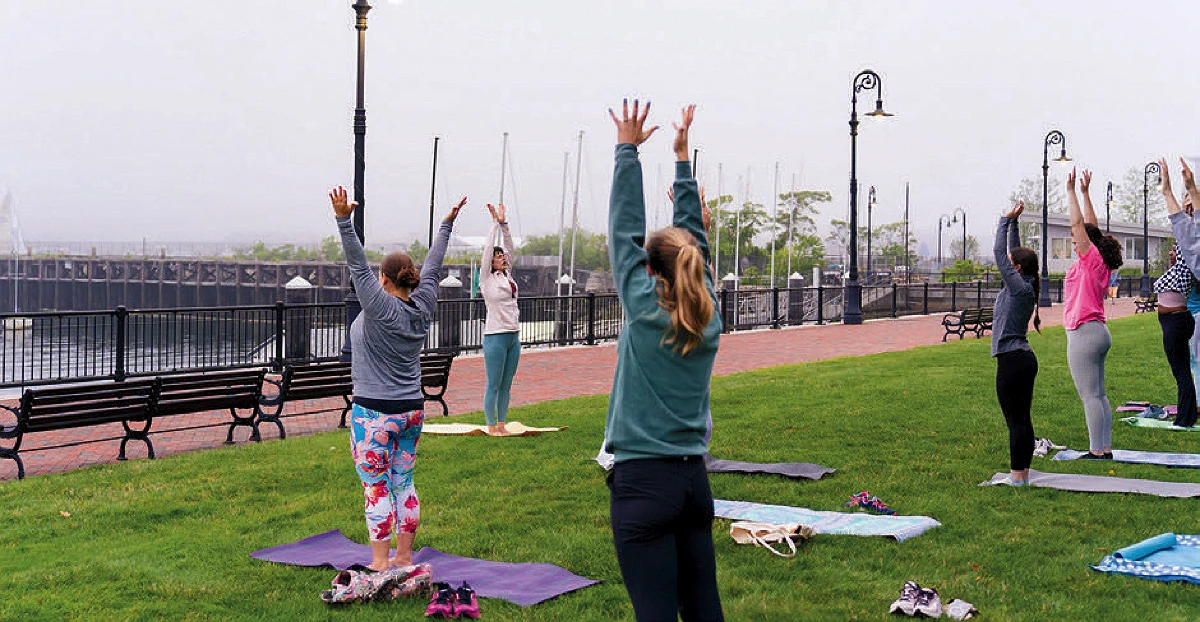 People practicing yoga on mats in a waterfront park, raising arms upward during a morning outdoor class.