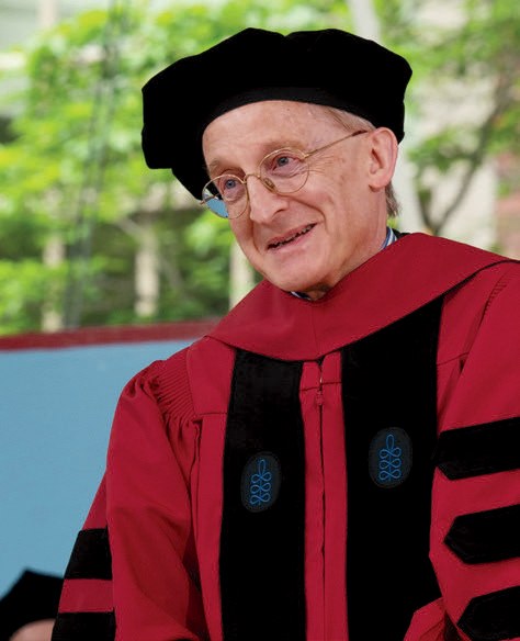 Person in red academic regalia and black doctoral cap smiles while speaking at commencement.