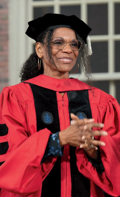 Person in red academic regalia and black doctoral cap gestures while addressing graduates outdoors.