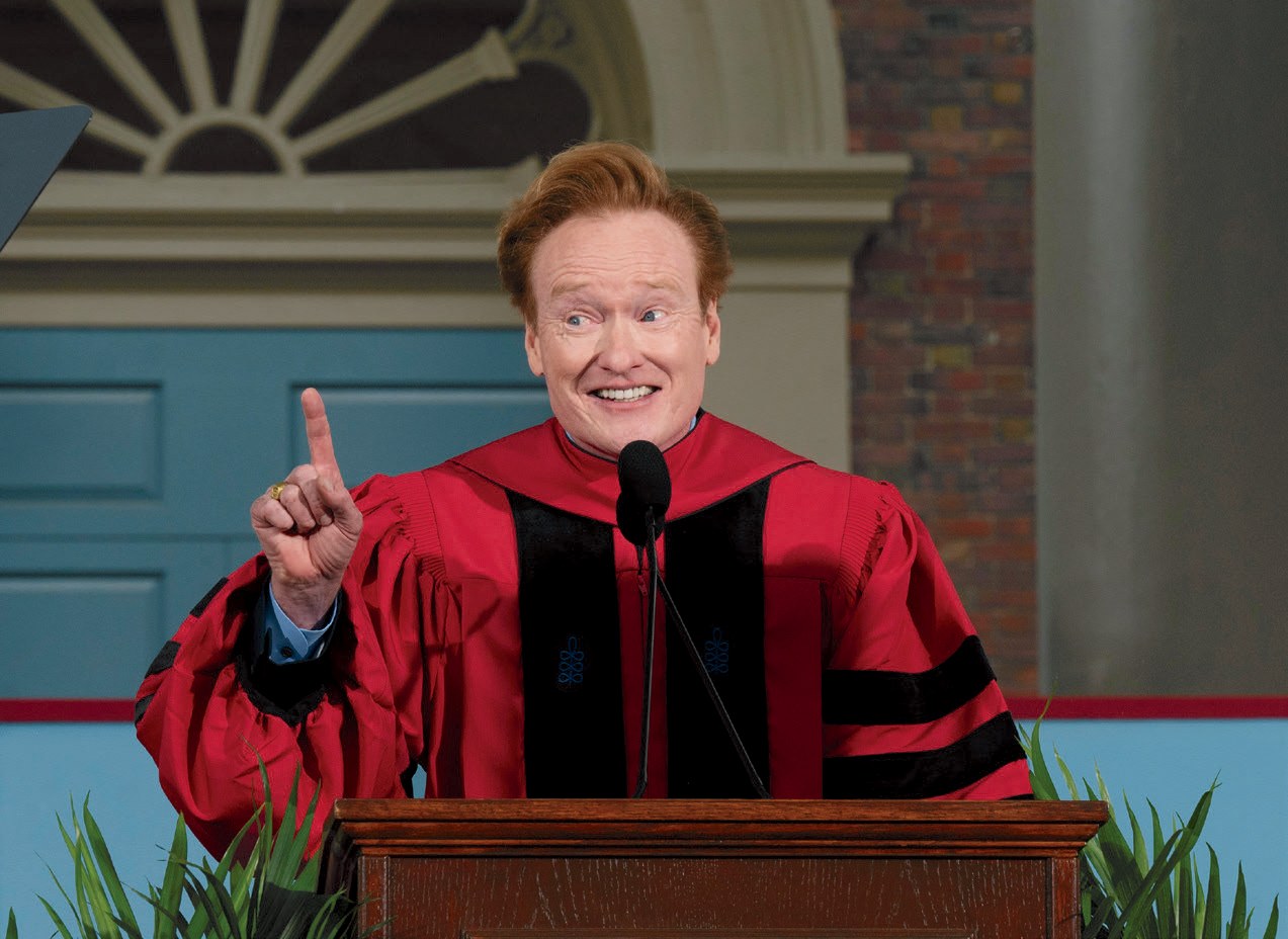 Conan O’Brien in red doctoral robe speak at a podium