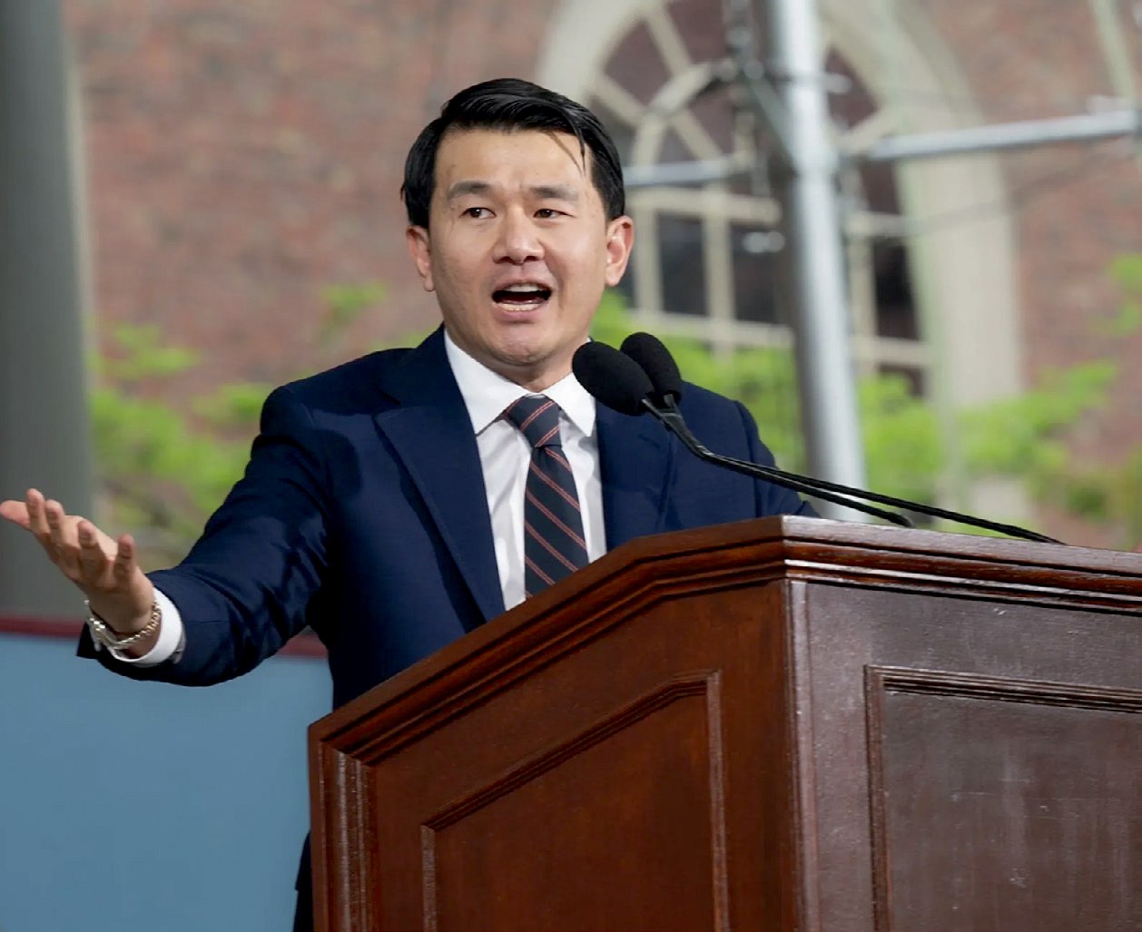 Ronny Chieng wearing a navy suit addresses audience from podium