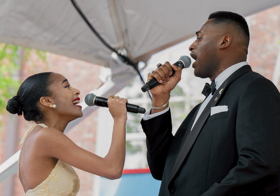 Two singers perform a duet with microphones on an outdoor commencement stage.