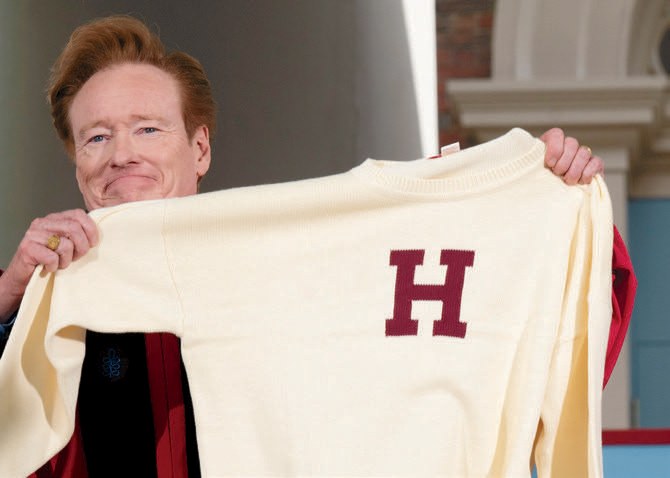 Person in academic regalia holds up a cream-colored sweater with a large crimson “H.”