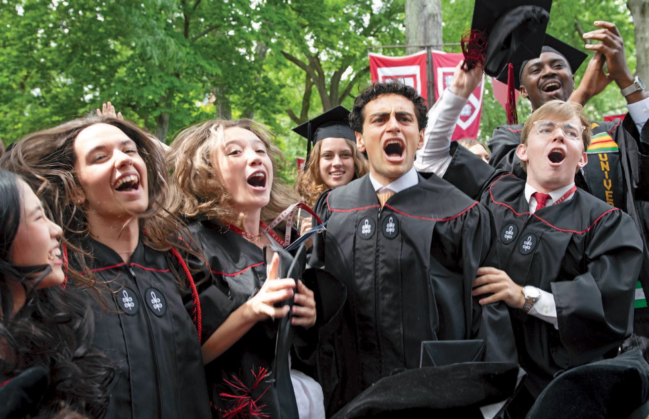 Graduates in black caps and gowns cheer and celebrate together outdoors after commencement.