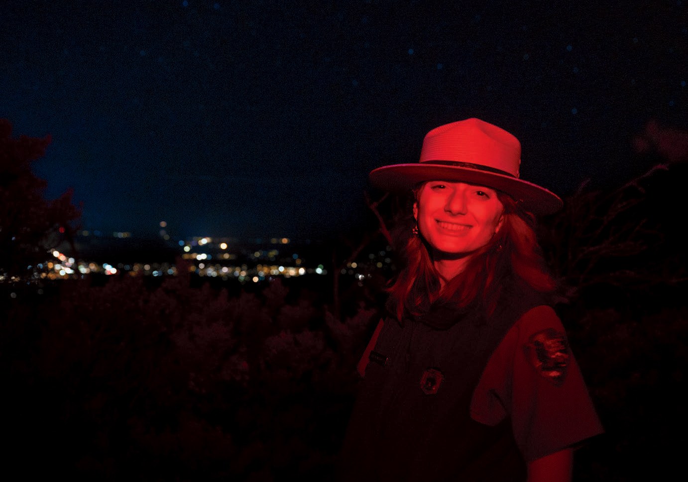 A woman wearing a wide brim hat smiling in red light in front of the night sky